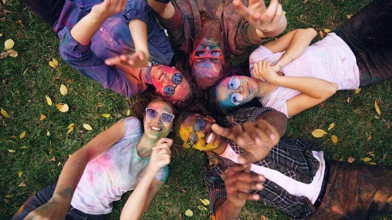 A group of happy friends with colorful face paint smiling and taking a selfie on a green grass field, representing the Promolyday lifestyle community.